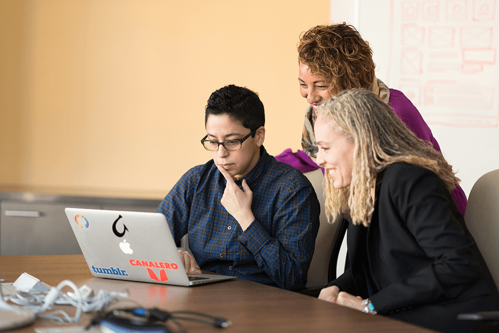 three women beside table looking at macbook